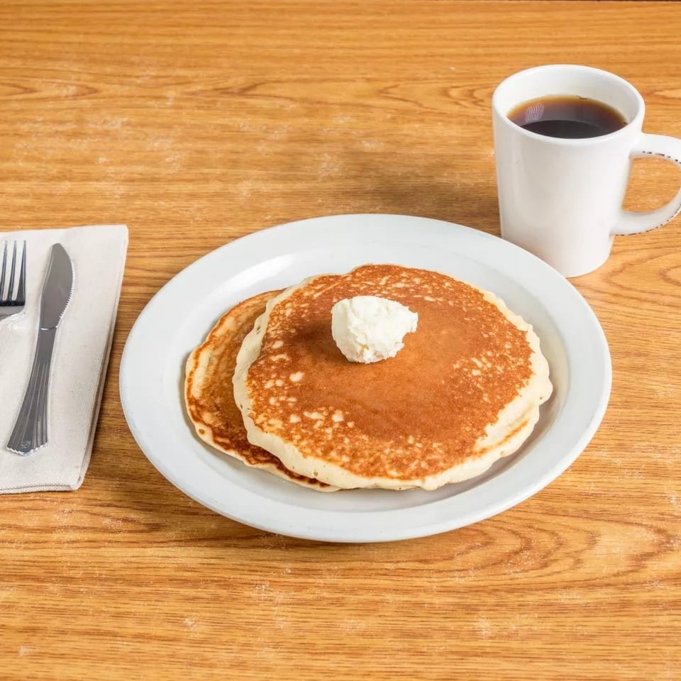 Two pancakes with butter, a cup of coffee, and silverware on a wooden table.