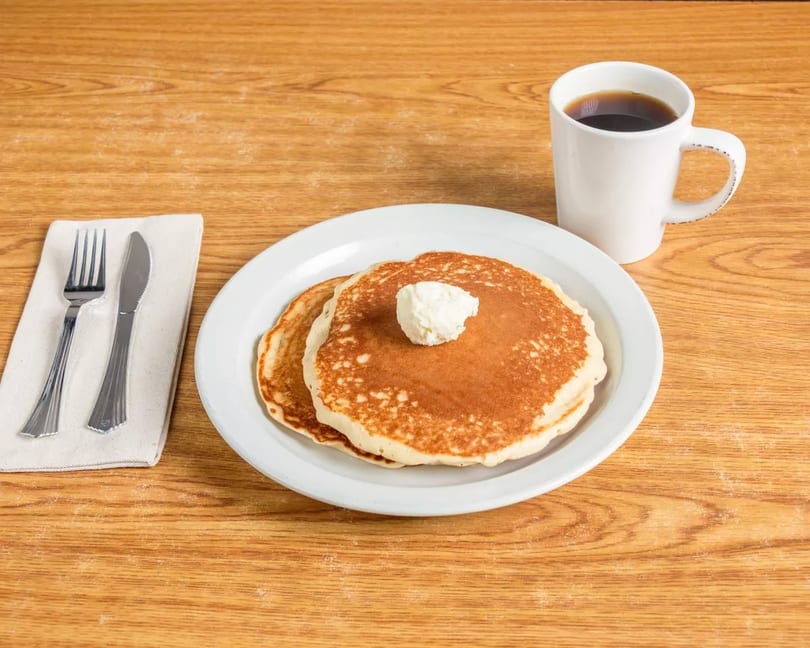 Two pancakes with butter, a cup of coffee, and silverware on a wooden table.