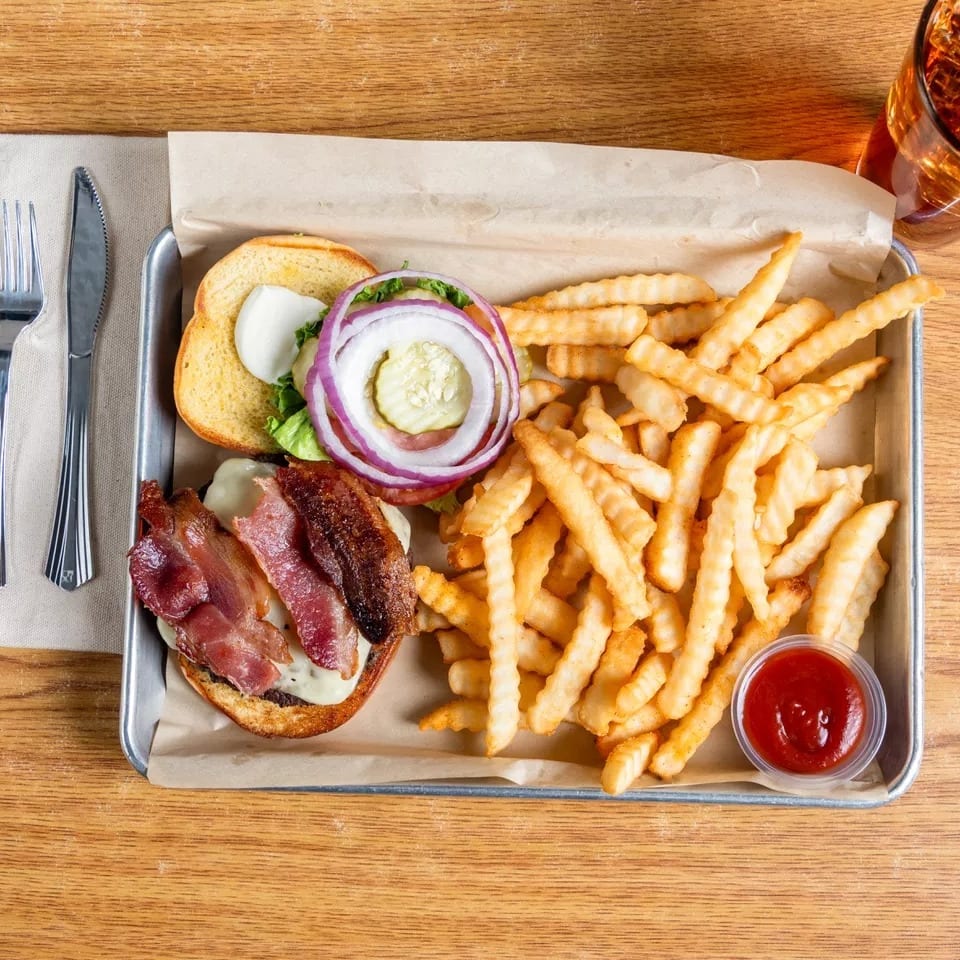 Bacon cheeseburger with fries and ketchup on a metal tray, with a drink and cutlery.
