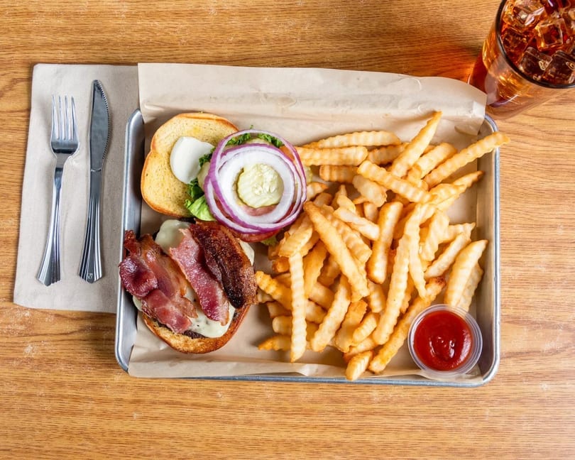 Bacon cheeseburger with fries and ketchup on a metal tray, with a drink and cutlery.