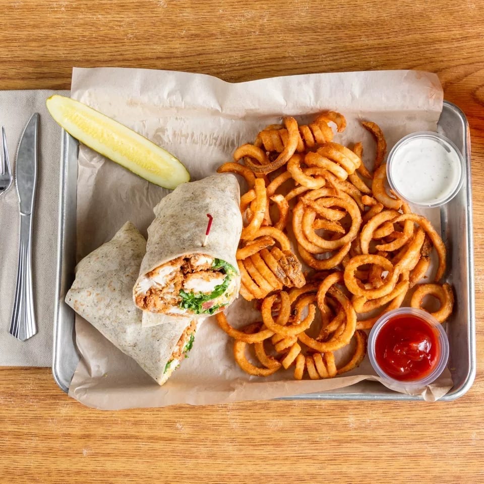 Chicken wrap with curly fries, a pickle spear, ketchup, and ranch dressing on a metal tray.