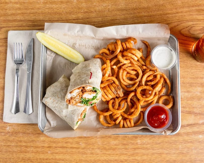 Chicken wrap with curly fries, a pickle spear, ketchup, and ranch dressing on a metal tray.