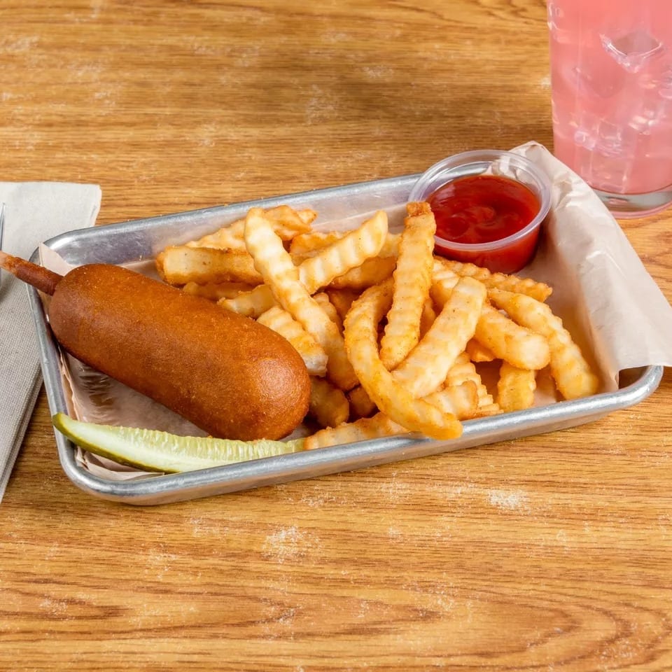 A corn dog, crinkle-cut fries, a pickle spear, and ketchup on a metal tray with pink lemonade.