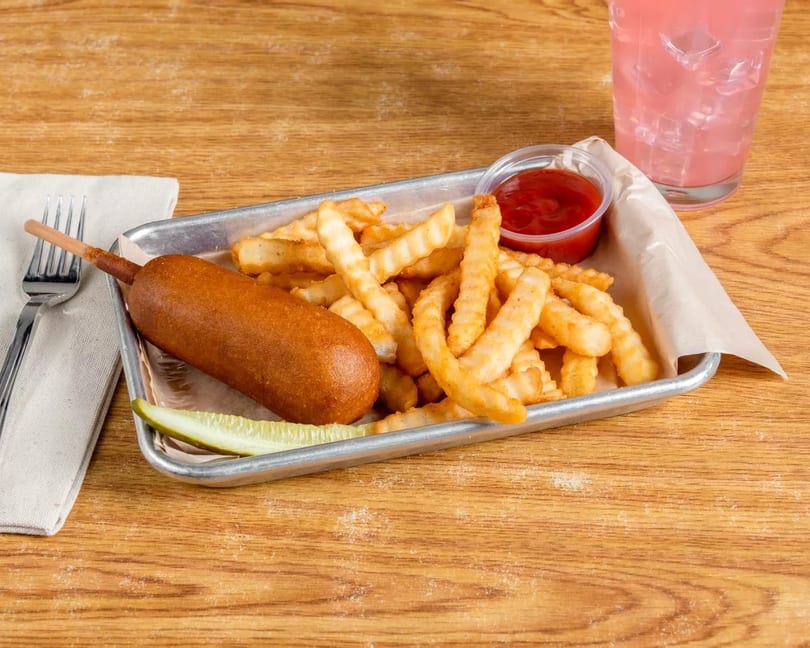 A corn dog, crinkle-cut fries, a pickle spear, and ketchup on a metal tray with pink lemonade.