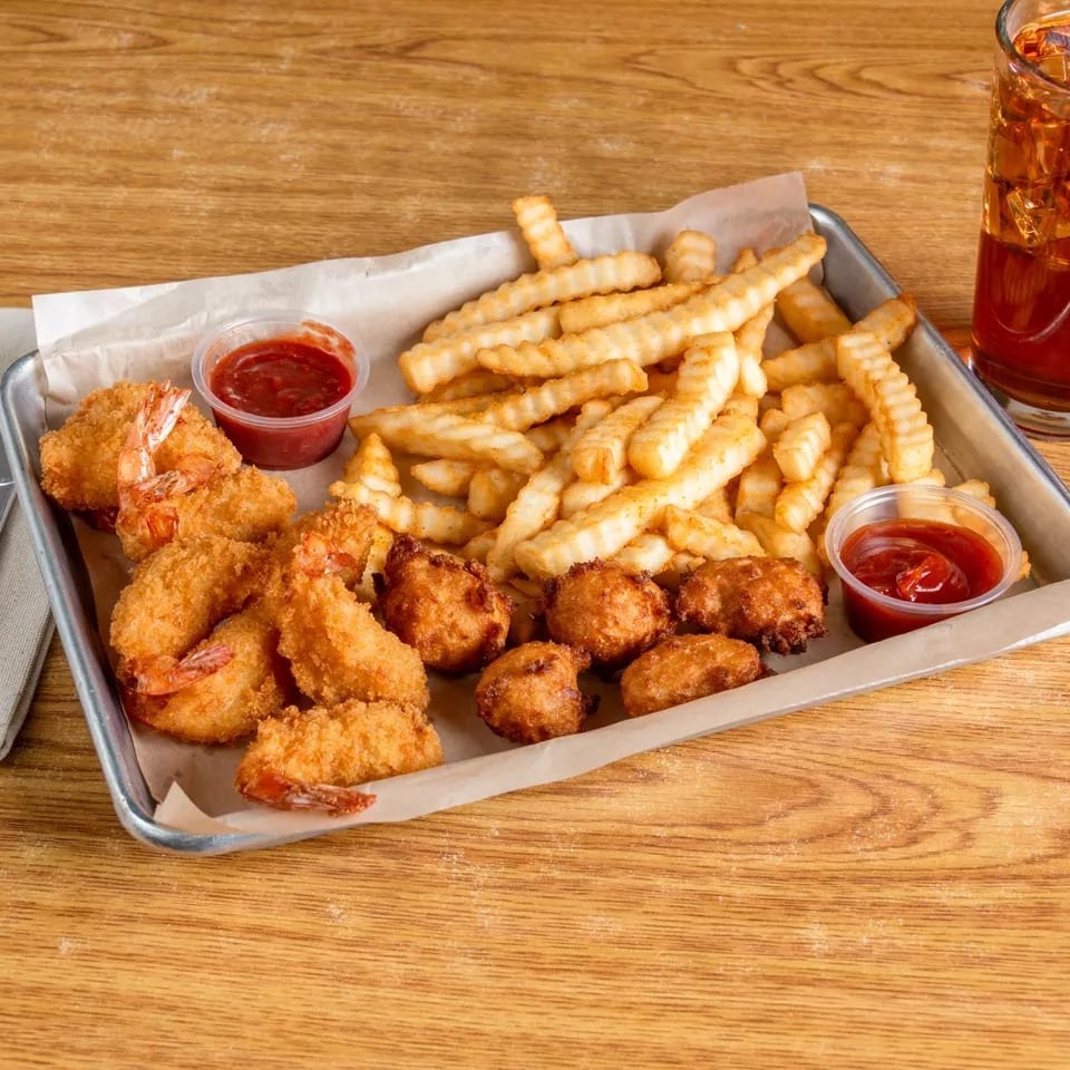 Fried shrimp, hush puppies, and crinkle-cut fries with ketchup and a drink on a wooden table.
