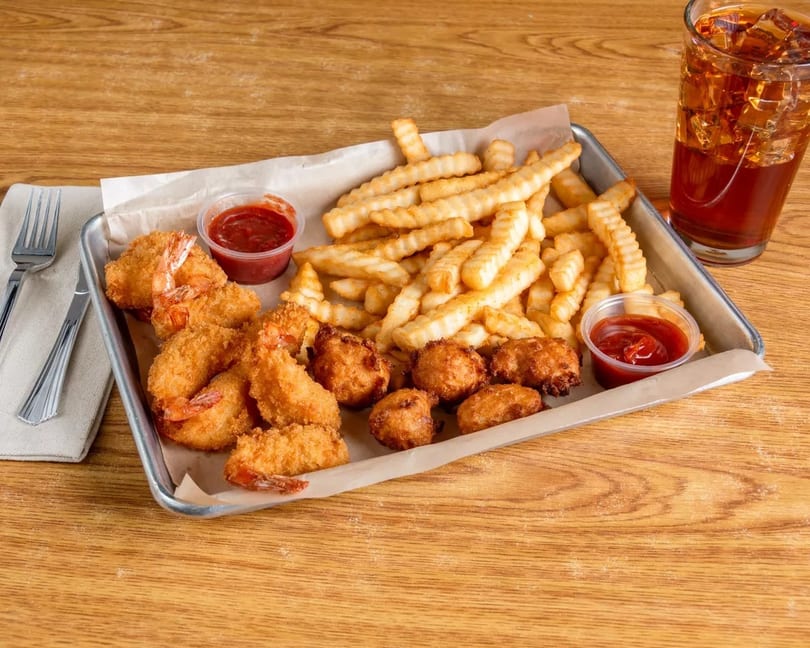 Fried shrimp, hush puppies, and crinkle-cut fries with ketchup and a drink on a wooden table.