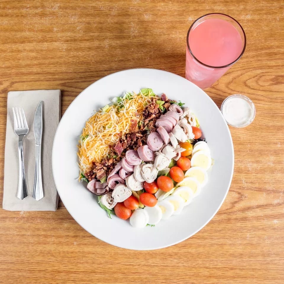 Overhead view of a large Cobb salad with a pink drink and dressing on a wooden table.
