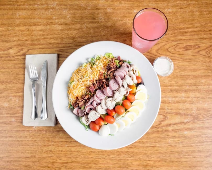 Overhead view of a large Cobb salad with a pink drink and dressing on a wooden table.