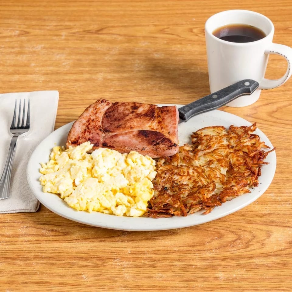 Breakfast plate with scrambled eggs, hash browns, ham steak, and a cup of coffee.