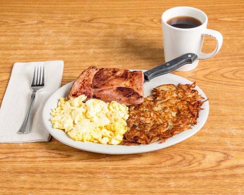Breakfast plate with scrambled eggs, hash browns, ham steak, and a cup of coffee.